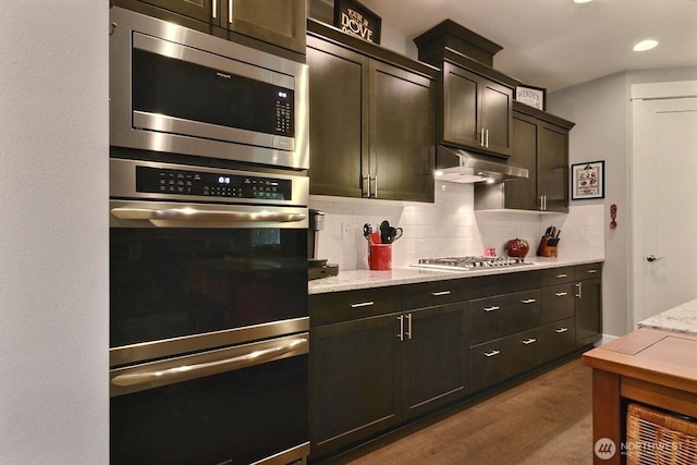 kitchen with light stone counters, decorative backsplash, dark wood-type flooring, appliances with stainless steel finishes, and under cabinet range hood