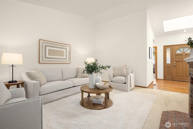 living room featuring high vaulted ceiling, light wood-style floors, a skylight, baseboards, and a brick fireplace