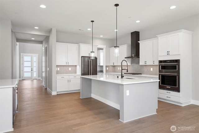 kitchen featuring a center island with sink, light wood finished floors, light countertops, appliances with stainless steel finishes, and wall chimney range hood