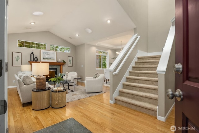 living area with wood finished floors, stairway, a lit fireplace, baseboards, and vaulted ceiling