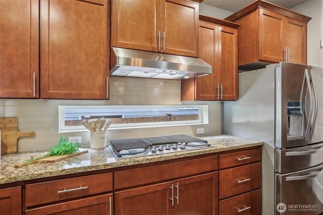 kitchen featuring under cabinet range hood, light stone counters, appliances with stainless steel finishes, brown cabinetry, and decorative backsplash