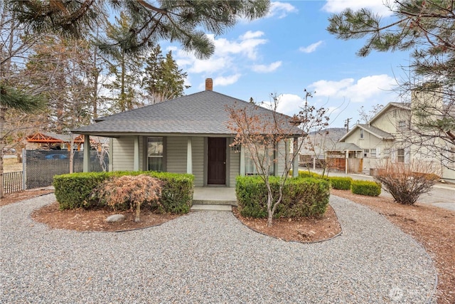 bungalow-style home featuring covered porch, a shingled roof, a chimney, and fence