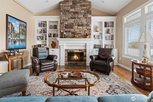 living area with baseboards, built in features, lofted ceiling, a stone fireplace, and wood finished floors