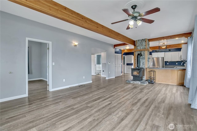 unfurnished living room with baseboards, beam ceiling, a wood stove, a sink, and light wood-style floors