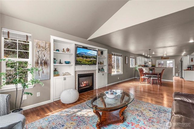 living room featuring built in shelves, wood finished floors, baseboards, vaulted ceiling, and a glass covered fireplace