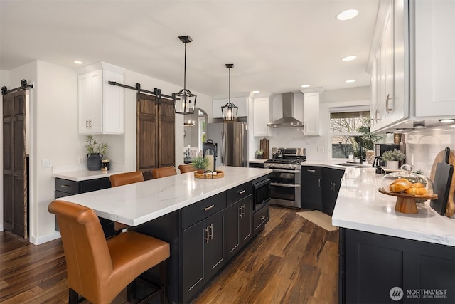 kitchen featuring white cabinetry, a barn door, stainless steel appliances, wall chimney range hood, and dark cabinets