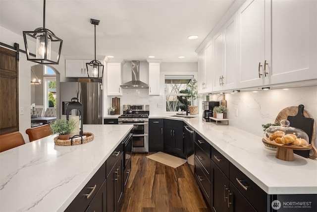 kitchen with stainless steel appliances, a barn door, wall chimney exhaust hood, and white cabinets