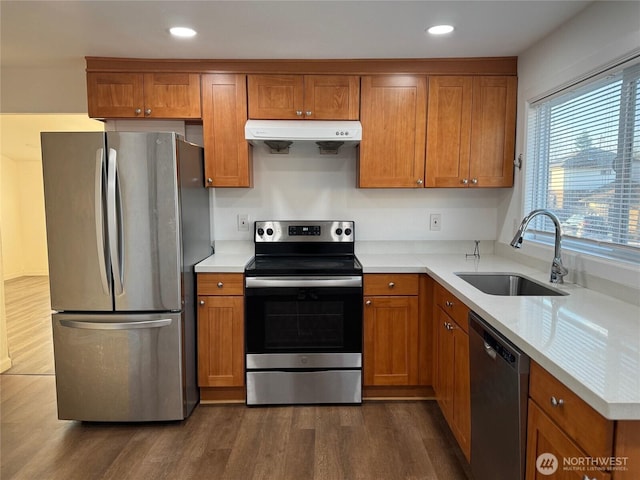 kitchen with a sink, under cabinet range hood, brown cabinetry, stainless steel appliances, and dark wood-style flooring