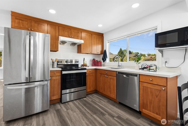 kitchen featuring brown cabinetry, a sink, light countertops, under cabinet range hood, and appliances with stainless steel finishes