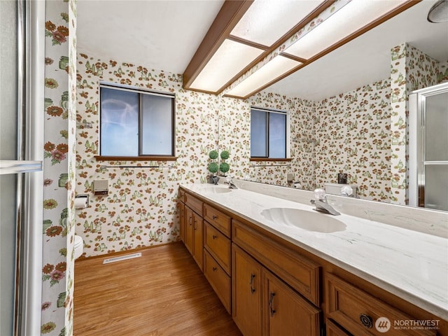 bathroom with double vanity, wood finished floors, wallpapered walls, and a sink