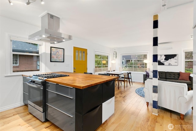 kitchen with dark cabinetry, island range hood, light wood-style flooring, stainless steel range with gas stovetop, and butcher block counters