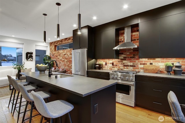 kitchen featuring a breakfast bar, high quality appliances, dark cabinetry, wall chimney exhaust hood, and a sink