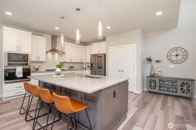 kitchen with a sink, tasteful backsplash, stainless steel appliances, light wood-style floors, and wall chimney exhaust hood