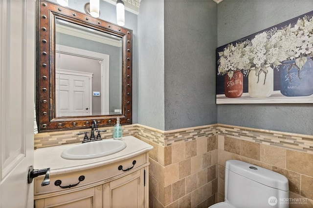 half bathroom featuring toilet, vanity, wainscoting, a textured wall, and tile walls