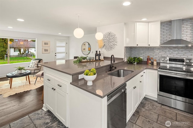 kitchen featuring a sink, white cabinetry, appliances with stainless steel finishes, a peninsula, and wall chimney range hood