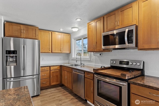 kitchen with brown cabinetry, light wood finished floors, a sink, stainless steel appliances, and a textured ceiling