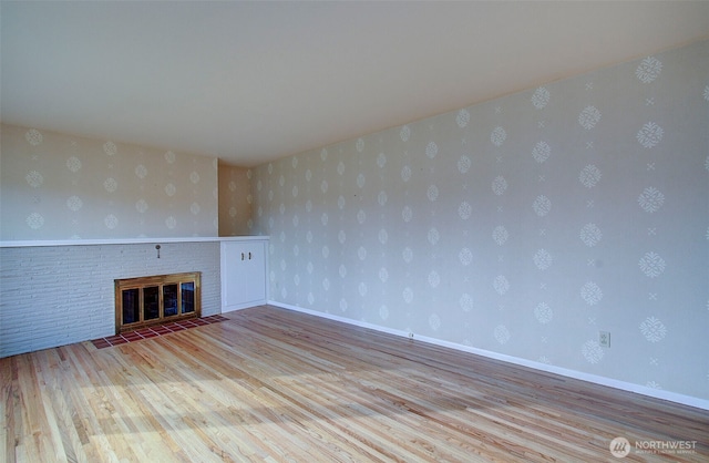 unfurnished living room with baseboards, a brick fireplace, wood finished floors, and wallpapered walls