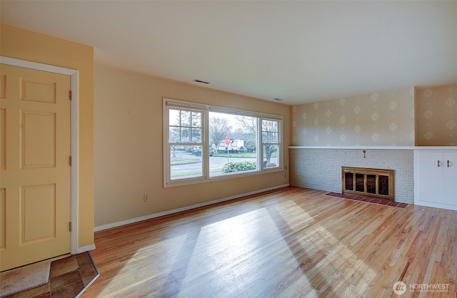 unfurnished living room with wallpapered walls, a brick fireplace, wood finished floors, and visible vents