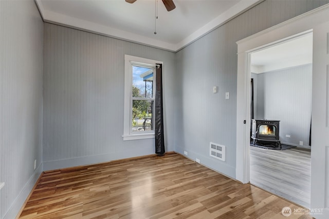 unfurnished room featuring ceiling fan, visible vents, wood finished floors, and a wood stove