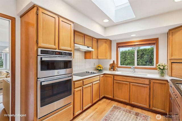 kitchen with a skylight, a sink, under cabinet range hood, double oven, and black electric cooktop