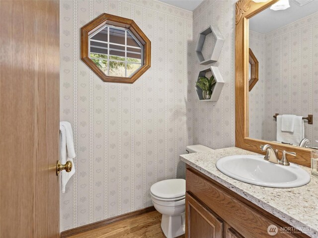 half bathroom with toilet, wallpapered walls, vanity, and wood finished floors