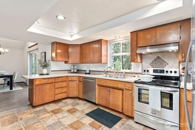 kitchen featuring under cabinet range hood, a tray ceiling, a sink, stainless steel appliances, and a peninsula