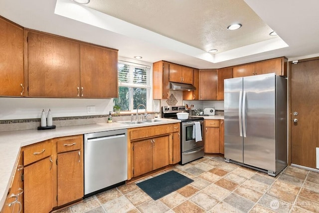 kitchen with a tray ceiling, a sink, stainless steel appliances, light countertops, and under cabinet range hood