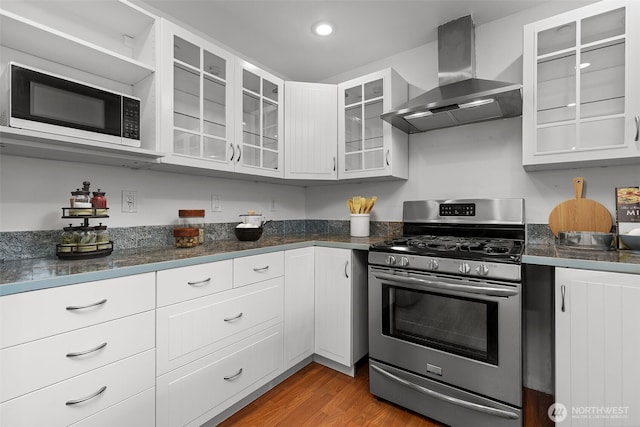 kitchen with glass insert cabinets, wall chimney range hood, gas range, wood finished floors, and white cabinets