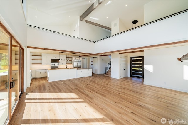 unfurnished living room featuring beam ceiling, stairway, light wood-style floors, and a towering ceiling