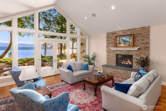 living room featuring a water view, high vaulted ceiling, wood finished floors, french doors, and a stone fireplace