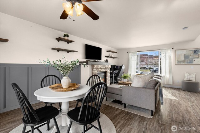 dining space featuring a ceiling fan, wood finished floors, a decorative wall, wainscoting, and a fireplace