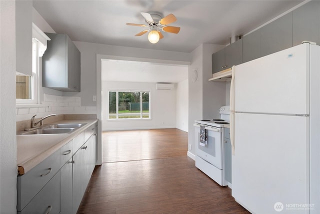 kitchen with a sink, gray cabinetry, a wall unit AC, white appliances, and dark wood-style flooring