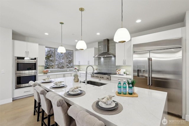 kitchen featuring a sink, backsplash, white cabinetry, appliances with stainless steel finishes, and wall chimney range hood