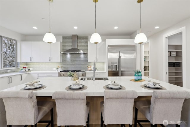 kitchen with a sink, built in fridge, white cabinetry, wall chimney exhaust hood, and light countertops