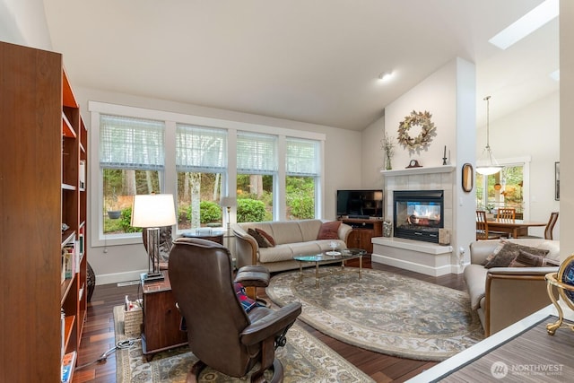 living area with dark wood finished floors, vaulted ceiling with skylight, a tiled fireplace, and baseboards