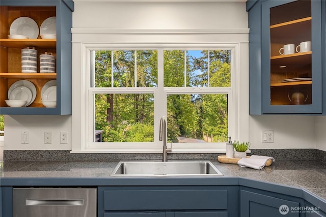 kitchen featuring a sink, tile counters, blue cabinetry, stainless steel dishwasher, and glass insert cabinets