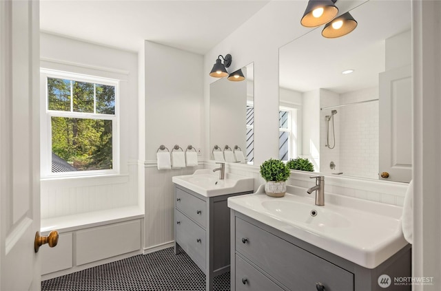 full bath featuring tile patterned flooring, wainscoting, two vanities, a sink, and tiled shower