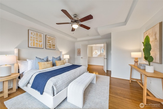 bedroom with ceiling fan with notable chandelier, a raised ceiling, wood finished floors, and baseboards