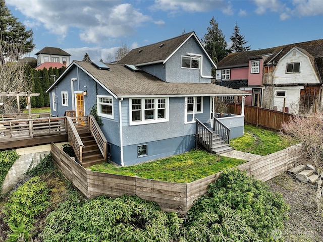 view of front of house with stucco siding, roof with shingles, a deck, and fence