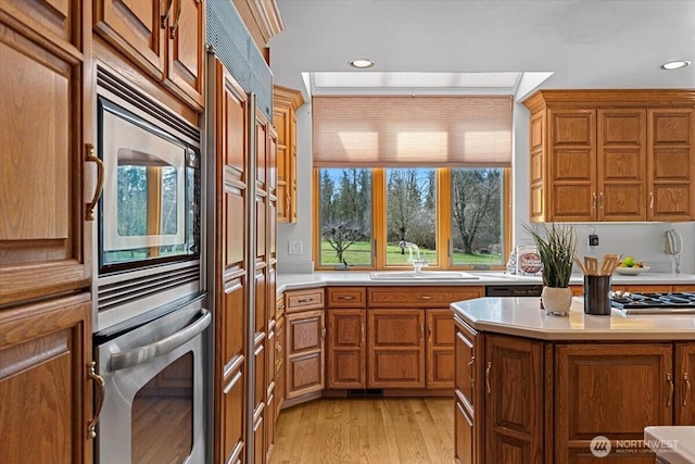 kitchen featuring a sink, stainless steel appliances, brown cabinetry, light wood finished floors, and light countertops