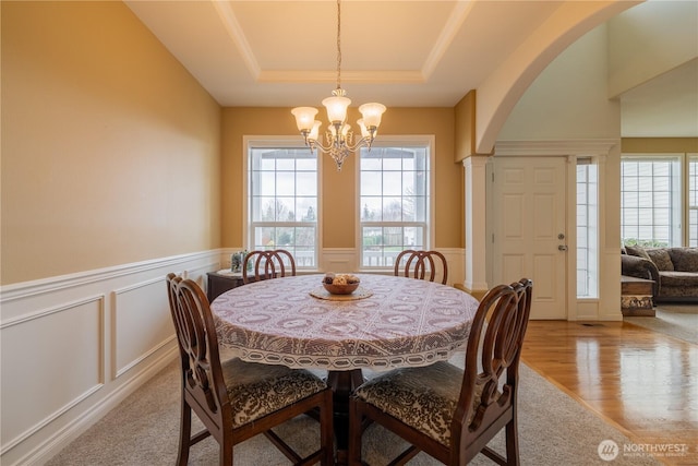 dining space with light wood-type flooring, a raised ceiling, arched walkways, and wainscoting