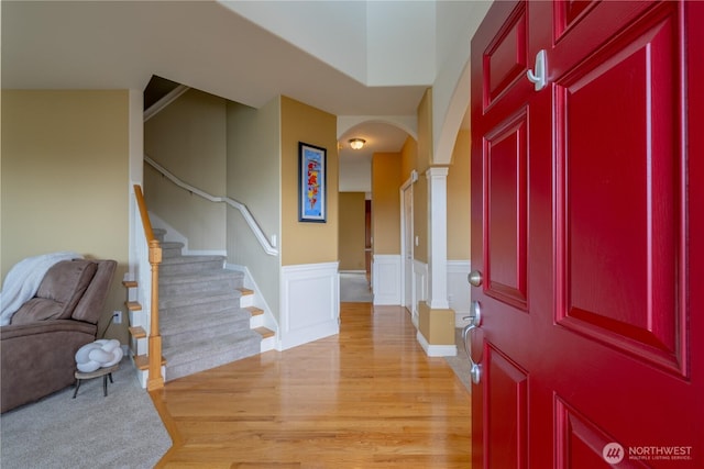 foyer entrance featuring a wainscoted wall, arched walkways, light wood-style floors, decorative columns, and stairs