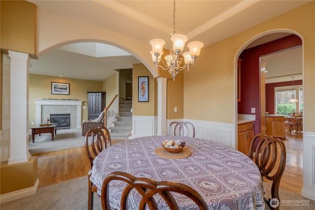 dining room with a tiled fireplace, a wainscoted wall, a chandelier, light wood-type flooring, and ornate columns