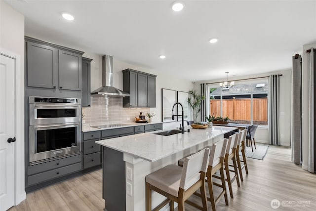 kitchen with a sink, double oven, wall chimney range hood, black electric stovetop, and light stone countertops