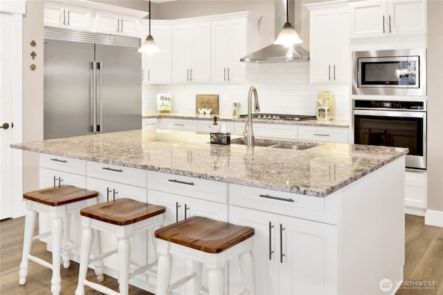 kitchen featuring backsplash, wall chimney range hood, built in appliances, light wood-style floors, and white cabinetry