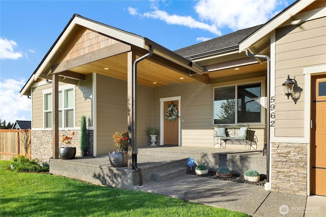 entrance to property with fence, covered porch, stone siding, and roof with shingles