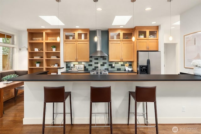 kitchen featuring dark countertops, wall chimney range hood, a skylight, and stainless steel fridge with ice dispenser