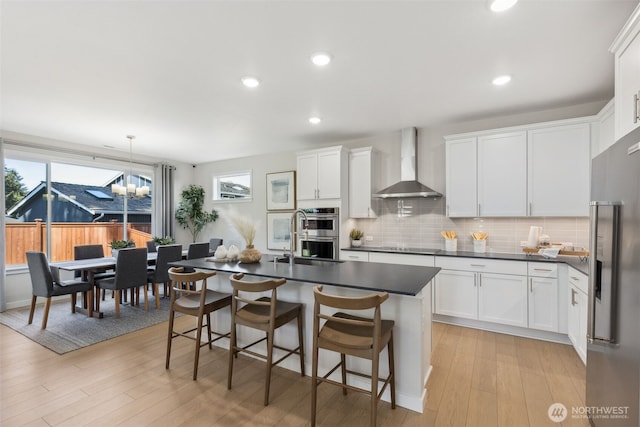 kitchen with light wood-type flooring, dark countertops, appliances with stainless steel finishes, wall chimney exhaust hood, and decorative backsplash