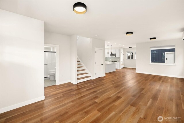 unfurnished living room featuring stairway, light wood-style flooring, and baseboards