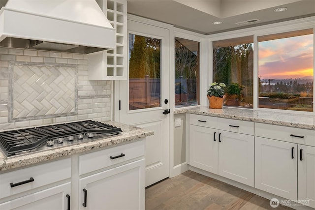 kitchen featuring light stone countertops, premium range hood, stainless steel gas stovetop, white cabinetry, and light wood-type flooring